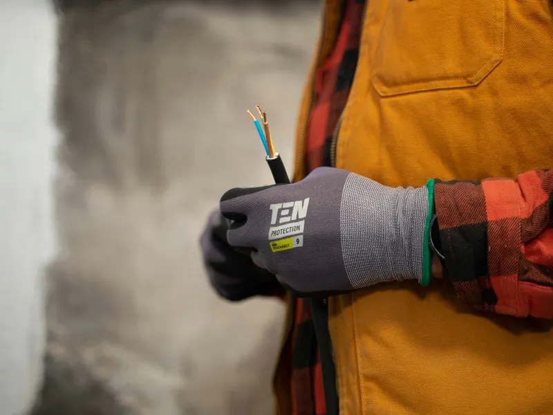 A gloved hand holding a stripped three-core cable, blue, brown and green-yellow conductors exposed, against an orange work jacket